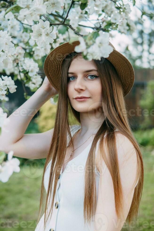 Beautiful young girl in white dress and hat in blooming Apple orchard. Blooming Apple trees with white flowers. photo