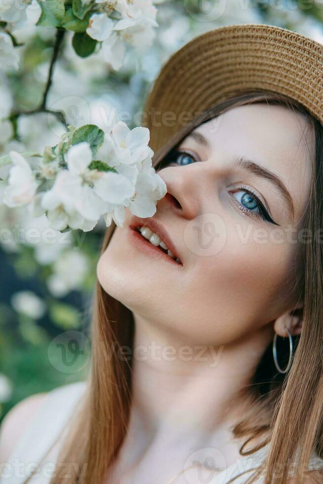 Beautiful young girl in white dress and hat in blooming Apple orchard. Blooming Apple trees with white flowers. photo