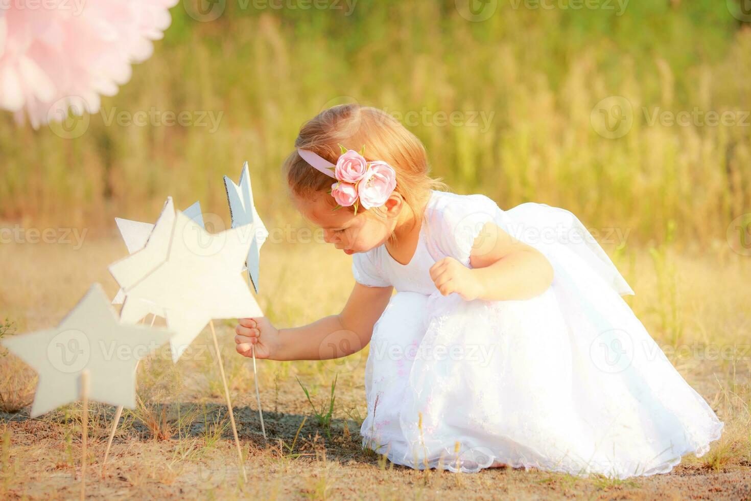 Beautiful girl in white long dress puts paper star on ground. Child on background of nature. photo