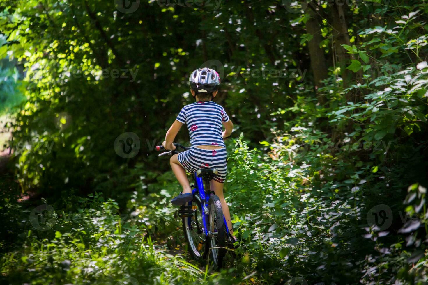 The little boy in a striped suit goes on a forest road. Child in a protective bicycle helmet riding a blue bike. photo