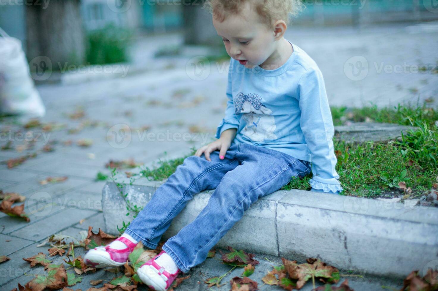 Cheerful little beautiful girl stands near a red wall. The child raised his hand walking down the street. photo