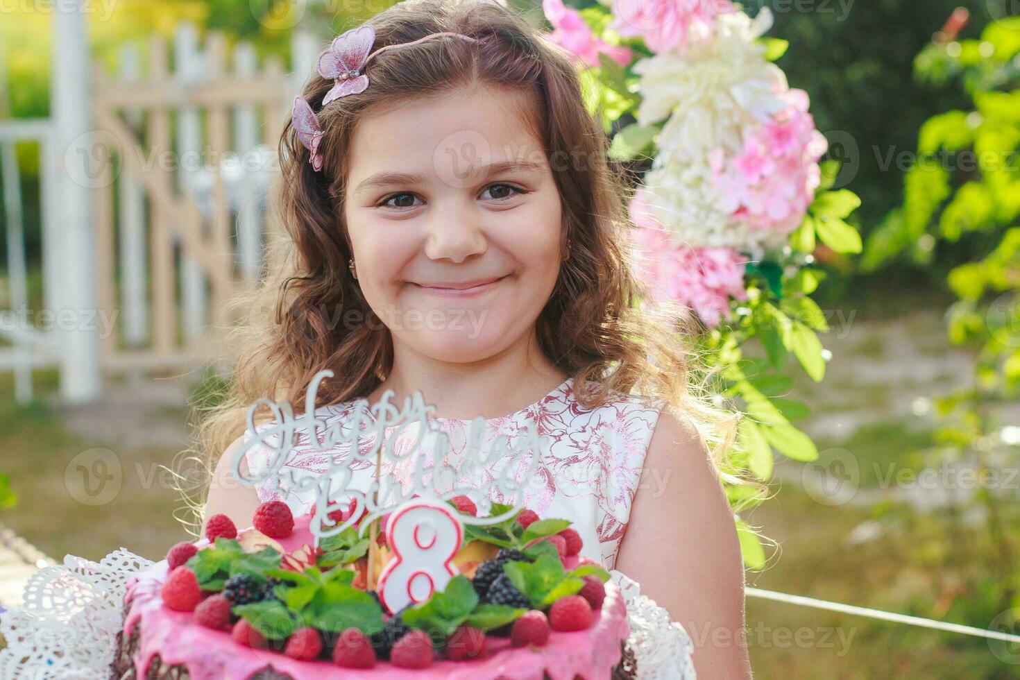 girl holding birthday cake with number 8 and inscription Happy Birthday. Celebrating ceremonial events on street during quarantine. child in background decorated photo zone of flowers.