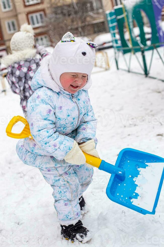 little girl with big shovel digs snow in the yard on playground. Active games outside house in winter during snowfall photo