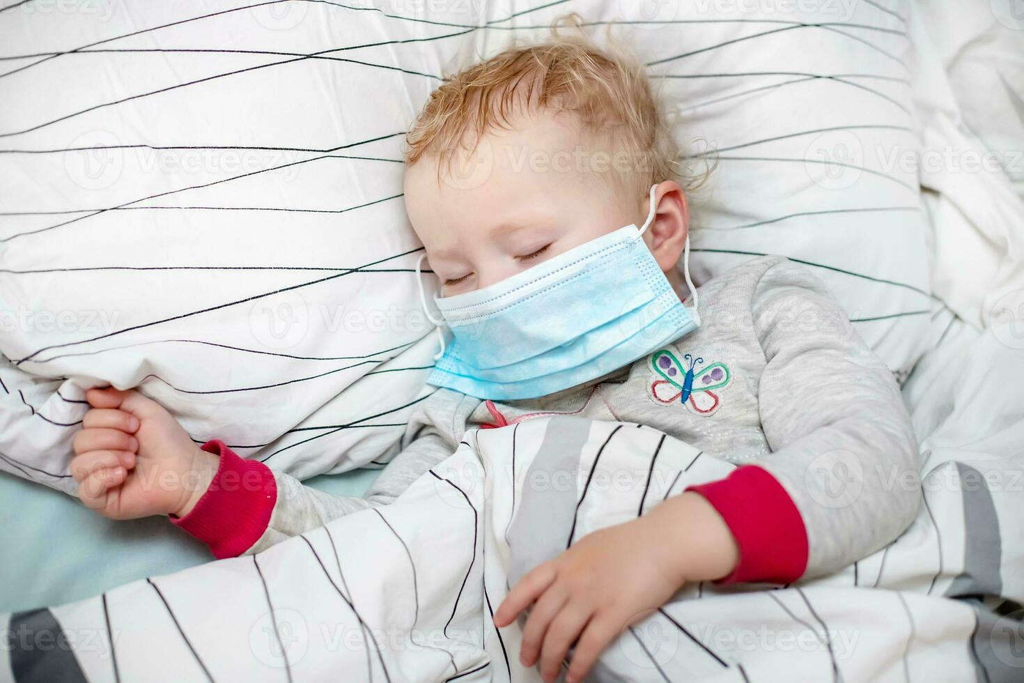 little child sleeps in bed in protective medical mask. sick child in white bed. Kid toddler one years old with flu, influenza or cold protected from viruses, pollution among patients with coronavirus photo