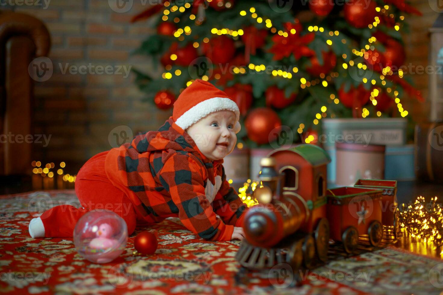 Little girl next to christmas train. Happy child in Santa hat near gifts on background of New Year tree in festive room. 6 month old baby is smiling. Beautiful little girl waiting for Christmas presents. photo