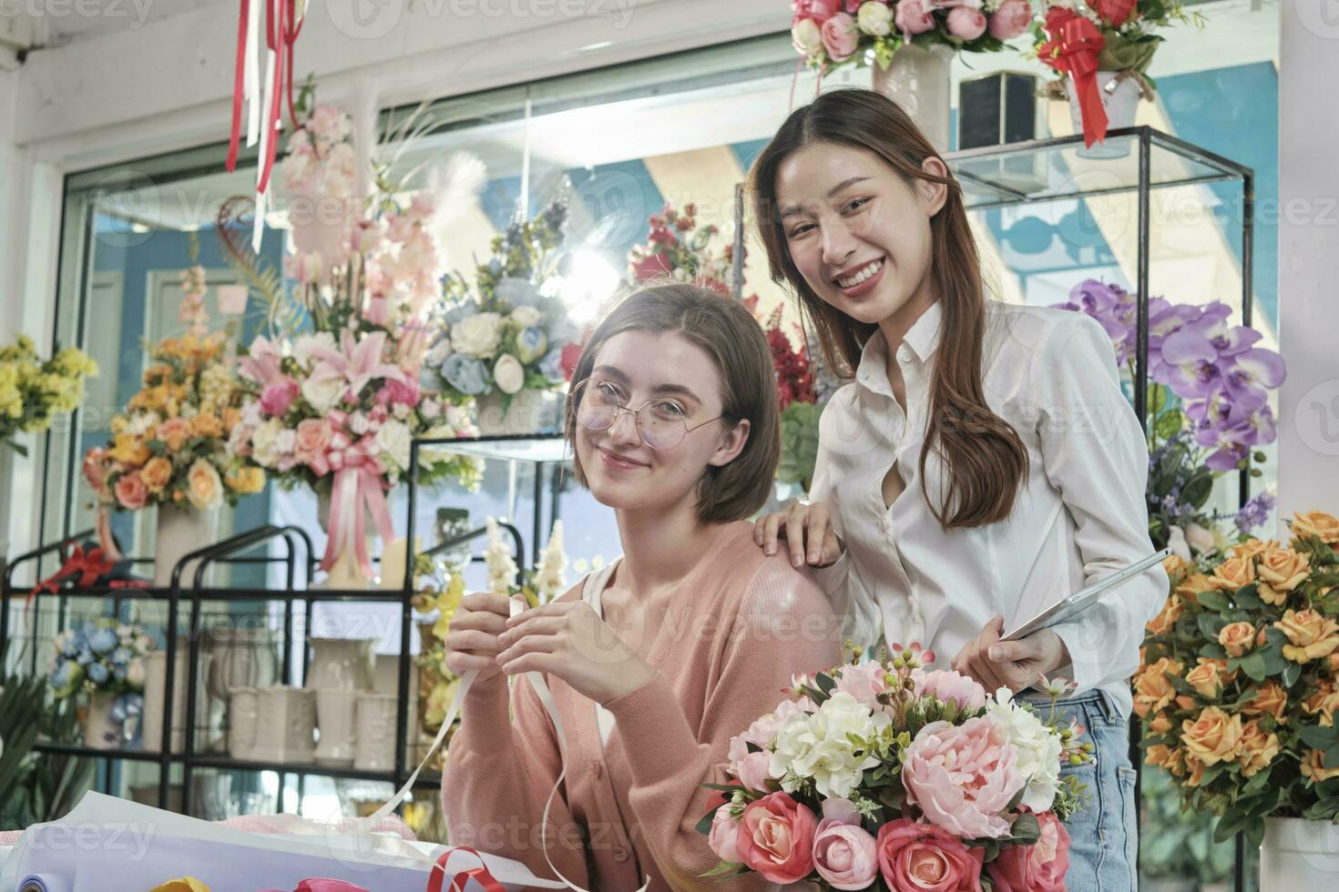 Portrait of two cheerful young beautiful female florist partners team smile and look at camera with bloom bouquet happy work in colorful flower shop store with fresh flora, SME business entrepreneur. photo