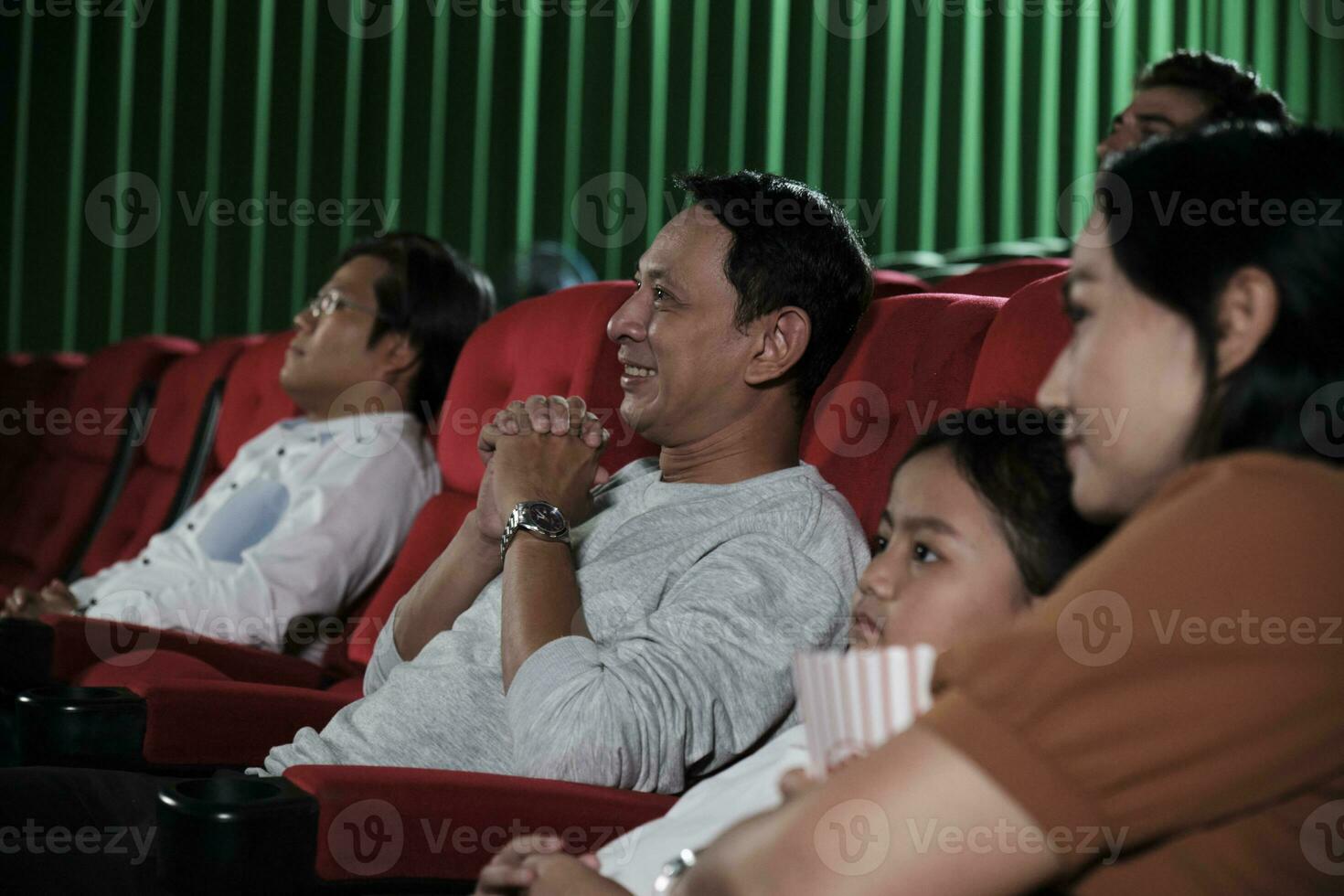 Asian family audience enjoys watching cinema together at movie theaters. Kid and parents have indoor entertainment lifestyle with performance art shows, happy and cheerful with popcorn and a smile. photo
