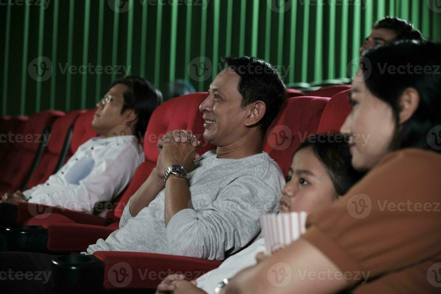 Asian family audience enjoys watching cinema together at movie theaters. Kid and parents have indoor entertainment lifestyle with performance art shows, happy and cheerful with popcorn and a smile. photo