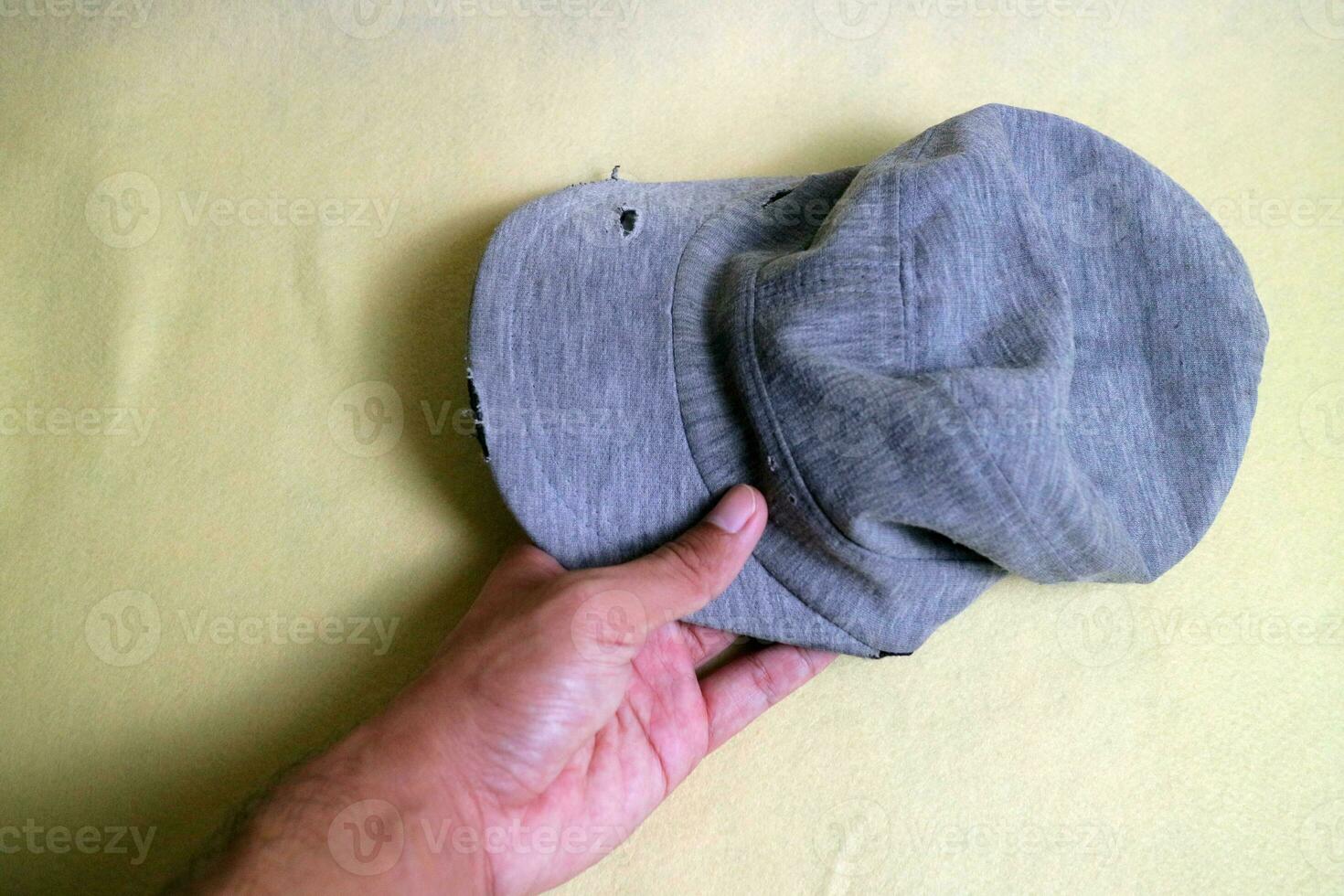 A hand holding a gray cloth hat is in a worn condition with several tears. Cream background photo