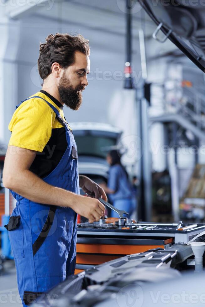 Serviceman in car service picks wrench from work station bench, using it to unscrew nuts in order to replace engine. Precise garage worker uses professional tools to fix customer automobile photo