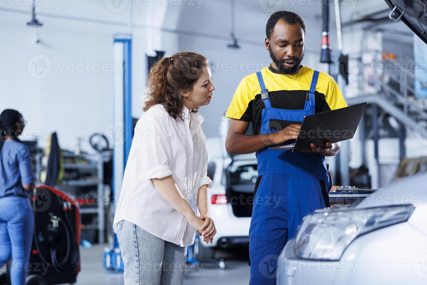 Repairman at auto repair shop conducts yearly vehicle checkup, informing customer about needed engine mending. BIPOC garage worker discussing with customer after finishing car maintenance photo