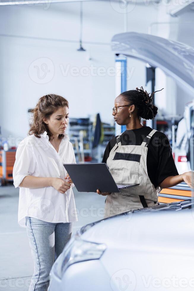Organized mechanic next to client in car service orders new engine for damaged vehicle using laptop. Employee looking for components on internet to replace old ones in faulty automobile photo
