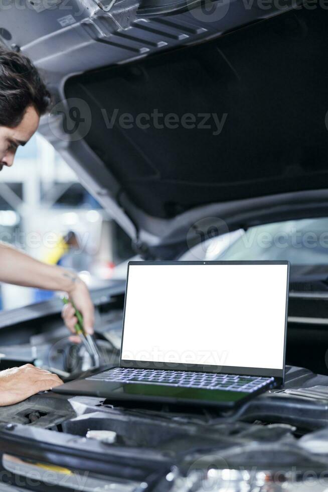 Mockup laptop on car with hood open while mechanic in blurry background replaces compressor belt. Isolated screen device next to skilled garage worker servicing client vehicle photo