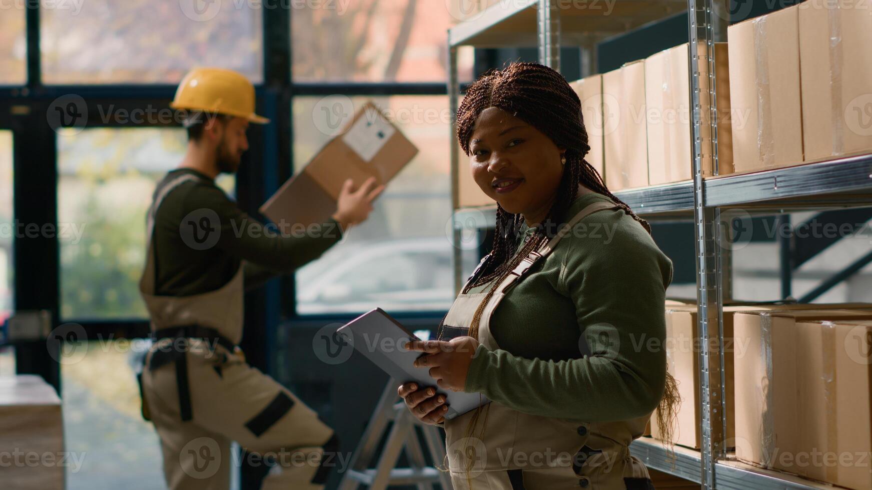 Portrait of joyful head of logistics in warehouse holding tablet, checking packaging while trainee in background handles packaged items in cardboard boxes ready to be shipped photo