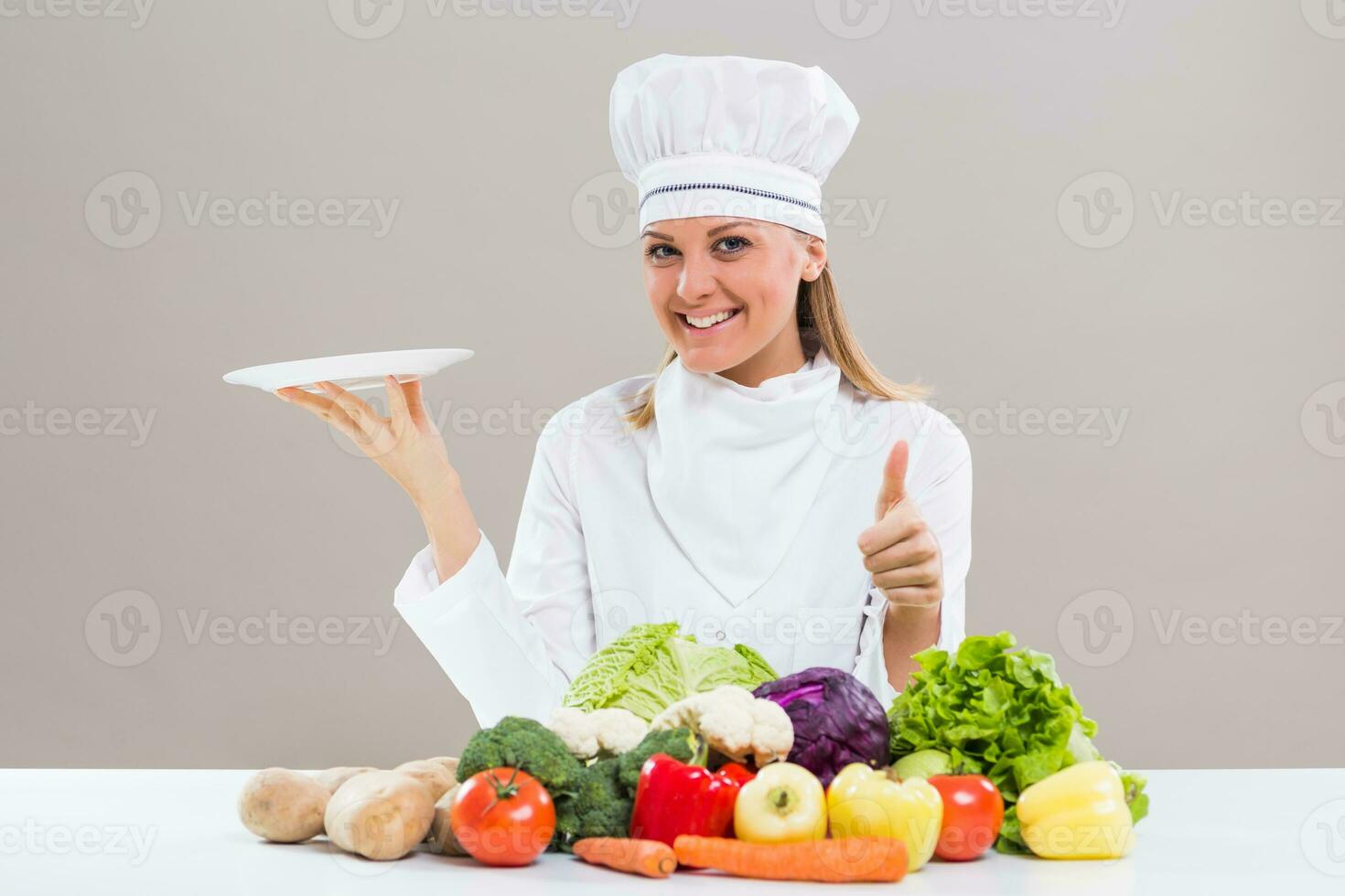 Female chef surrounded with vegetables holding plate showing thumb up photo