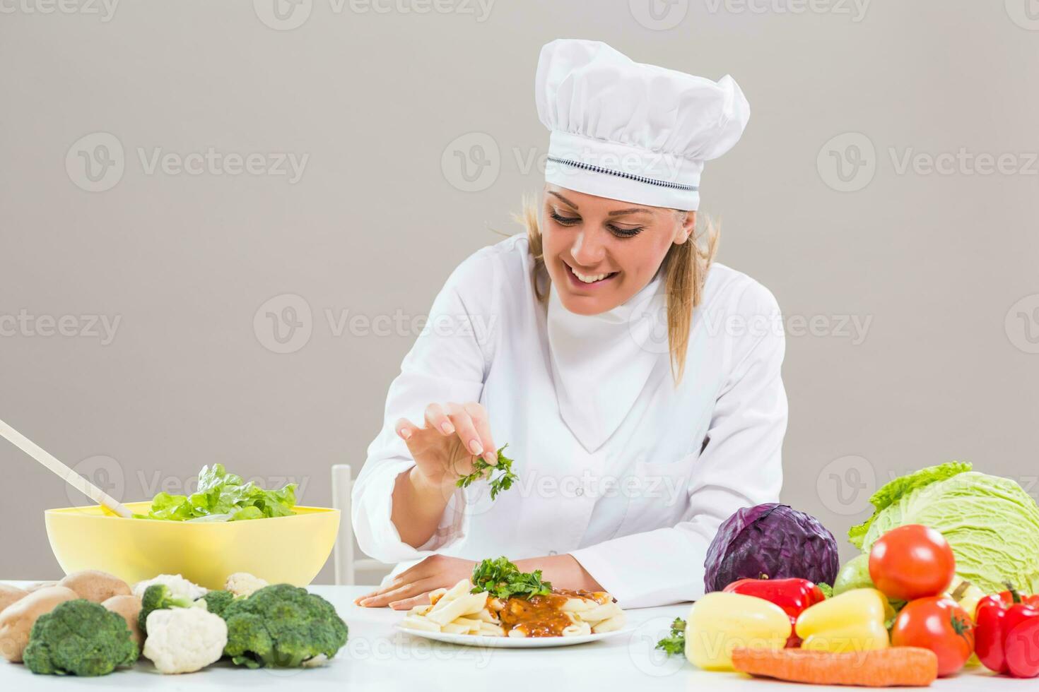 Cheerful female chef is sitting at the table with bunch of vegetable and decorating prepared meal. photo