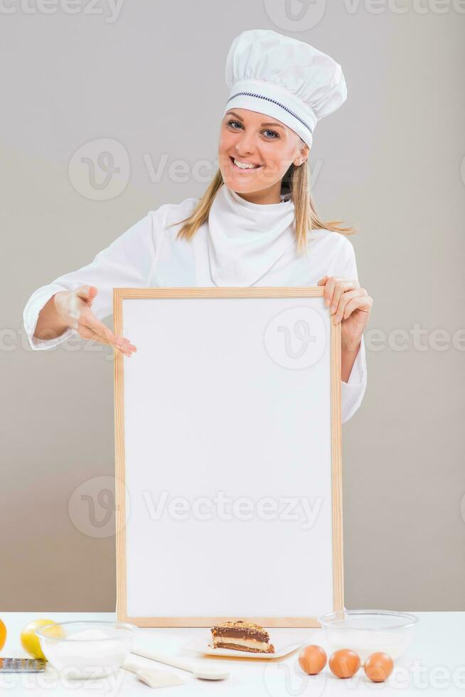 Beautiful female confectioner is showing empty whiteboard with slice of cake and ingredients for cake at the table. photo
