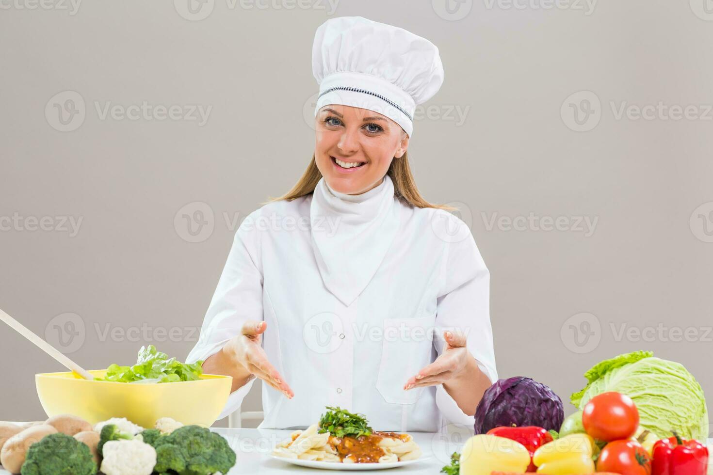 Content female chef is sitting at the table with bunch of vegetable showing prepared meal. photo