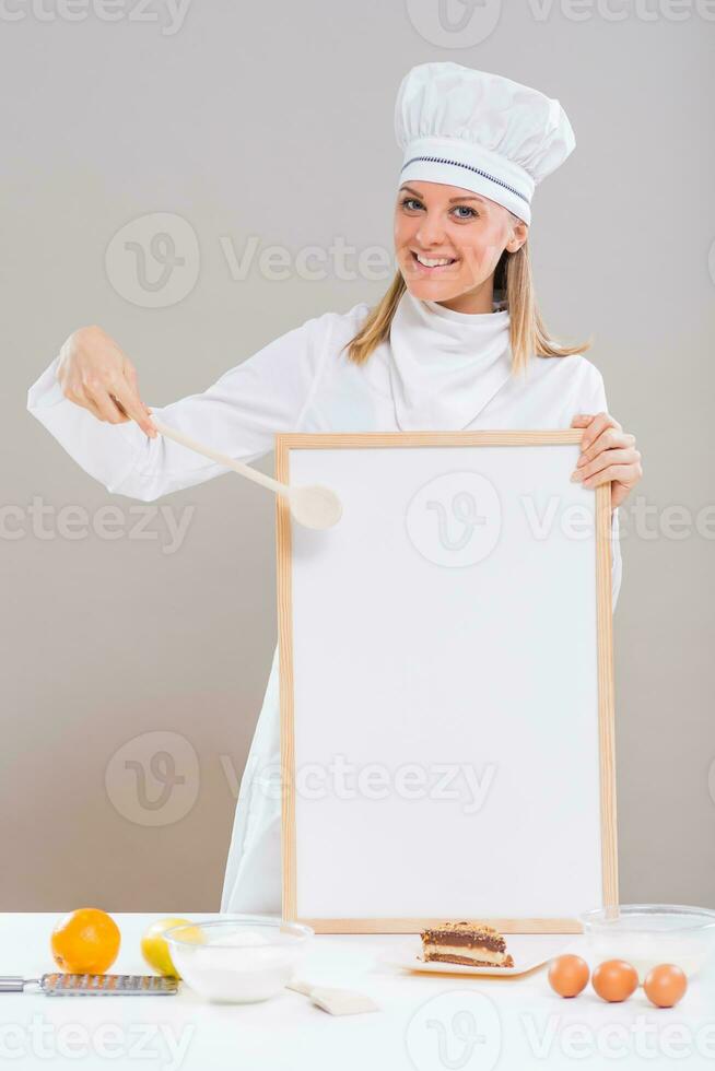 Beautiful female confectioner is showing whiteboard with slice of cake and ingredients for cake at the table. photo