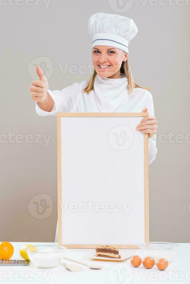 Beautiful female confectioner is showing thumb up while holding whiteboard with slice of cake and ingredients for cake at the table photo