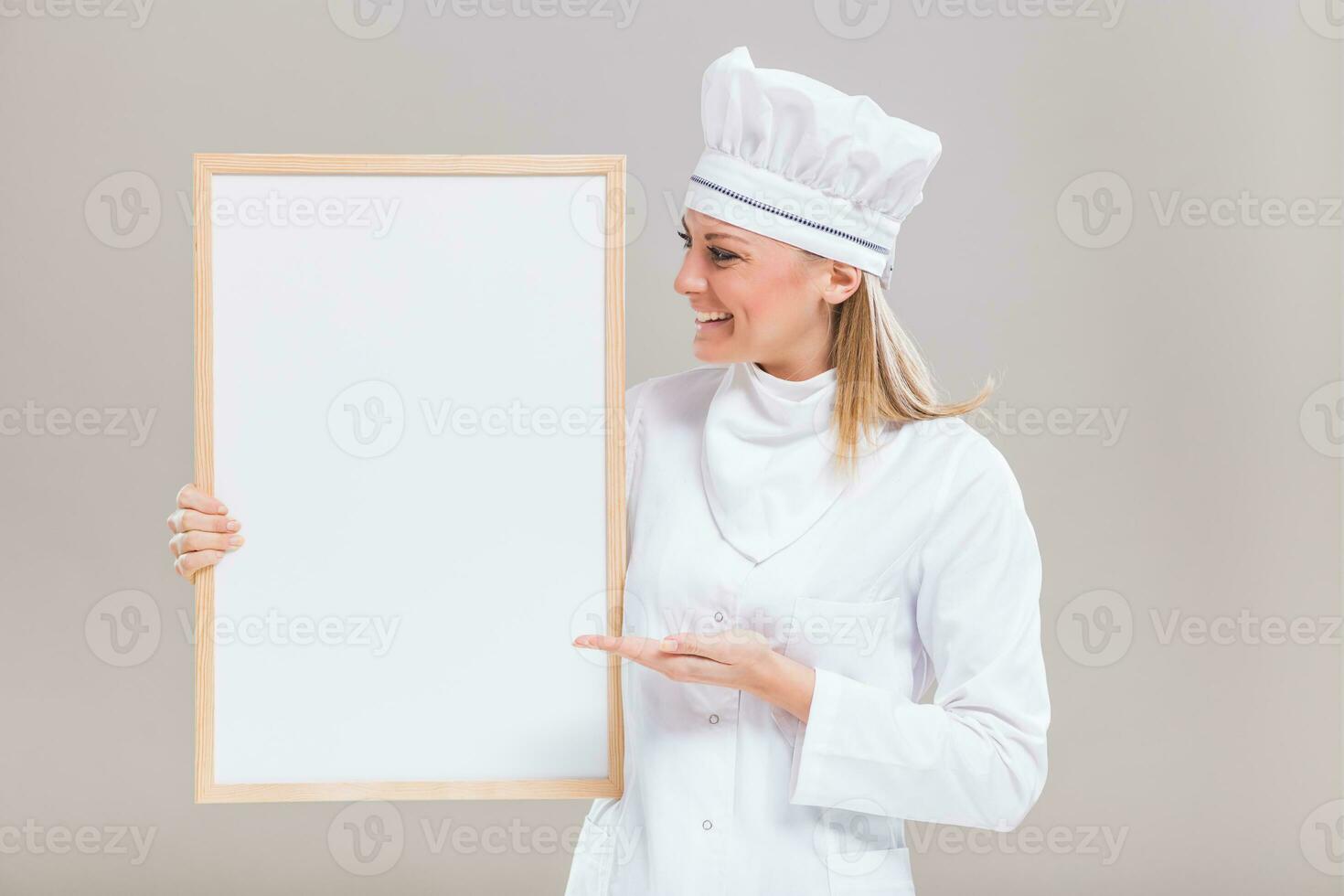 Portrait of beautiful female chef showing empty whiteboard on gray background. photo