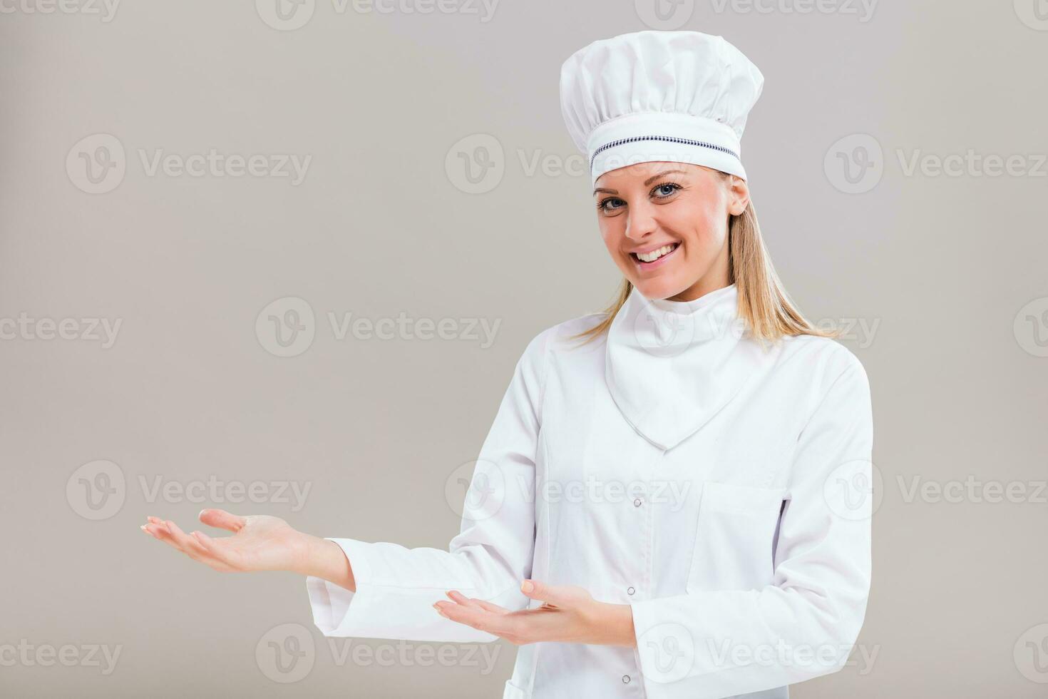 Portrait of beautiful female chef showing welcome gesture on gray background. photo