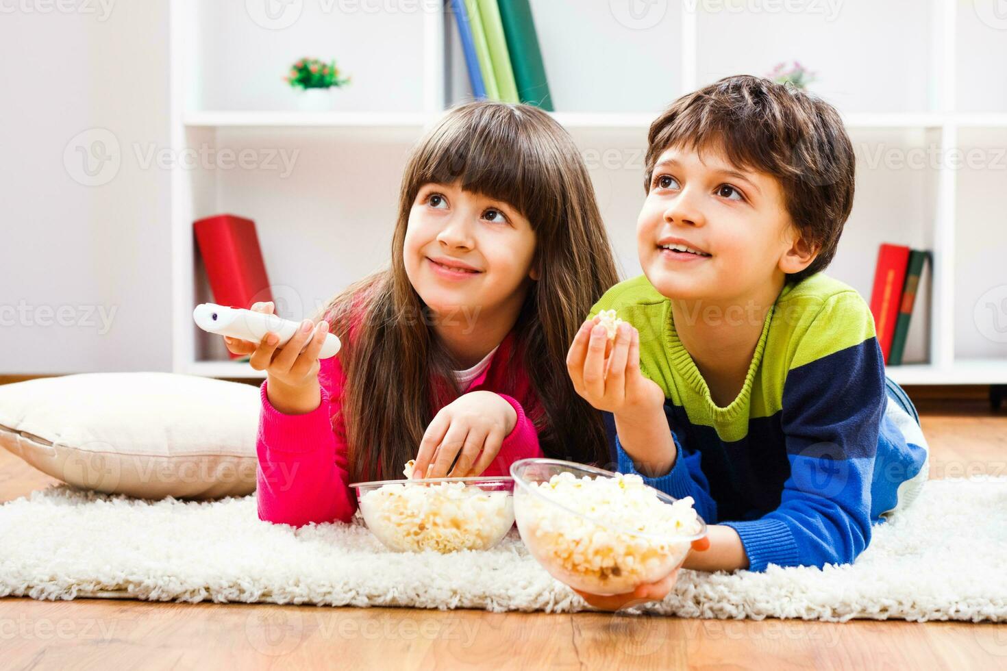 Two children are laying on the floor with popcorn and watching tv photo