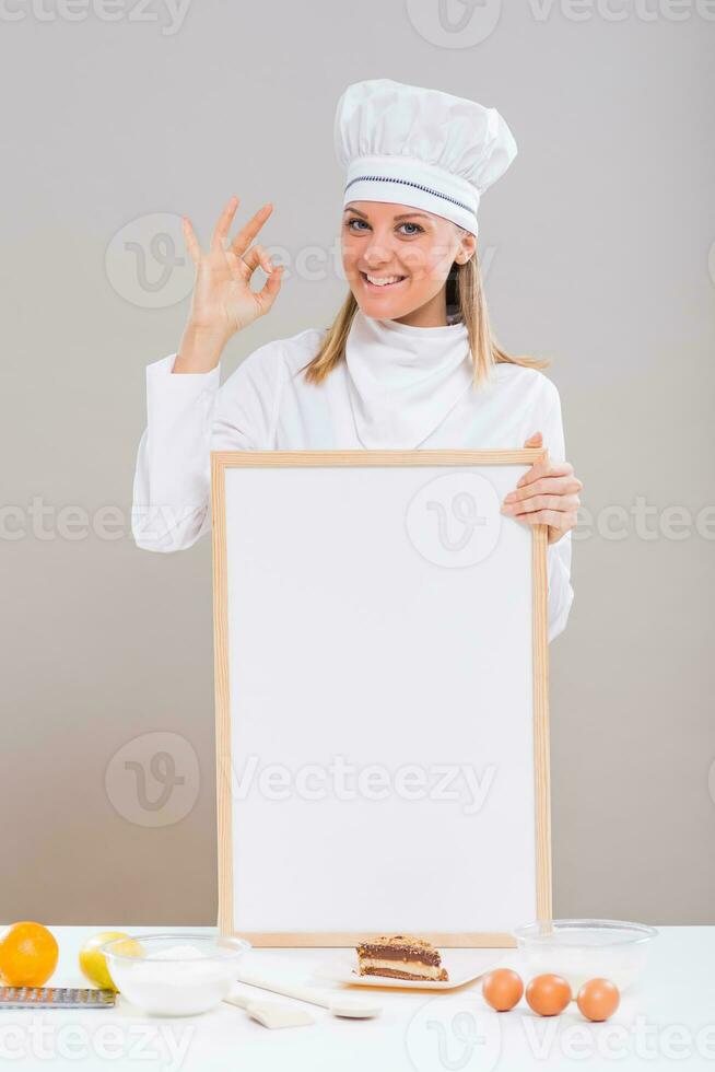 Beautiful female confectioner is showing ok sign while holding whiteboard with slice of cake and ingredients for cake at the table photo
