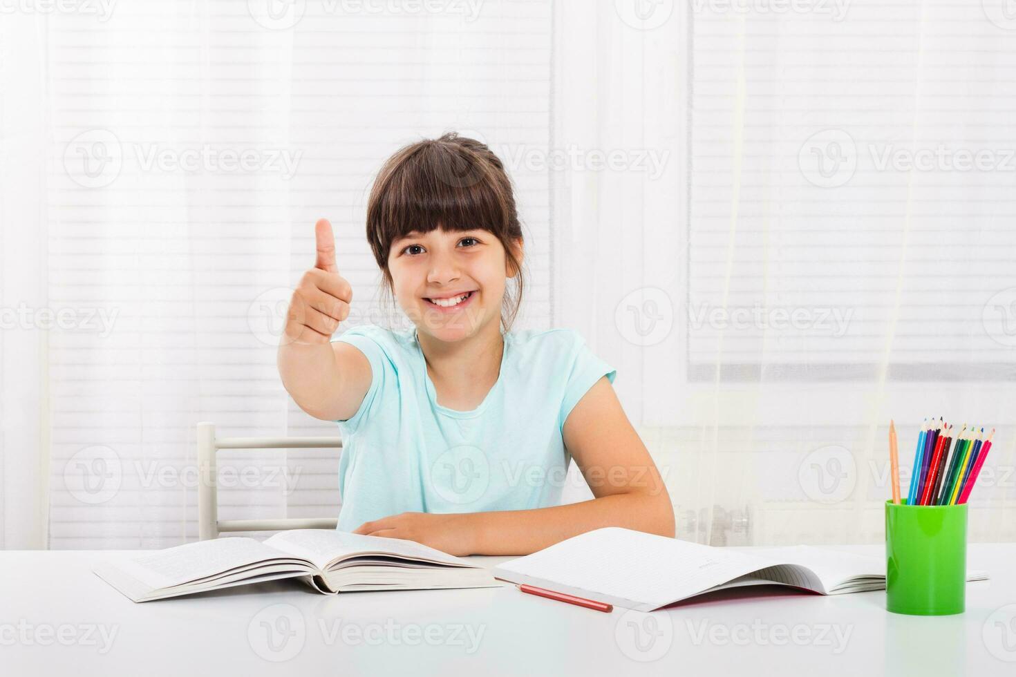 Cute little girl is showing thumbs up while she is doing her homework photo