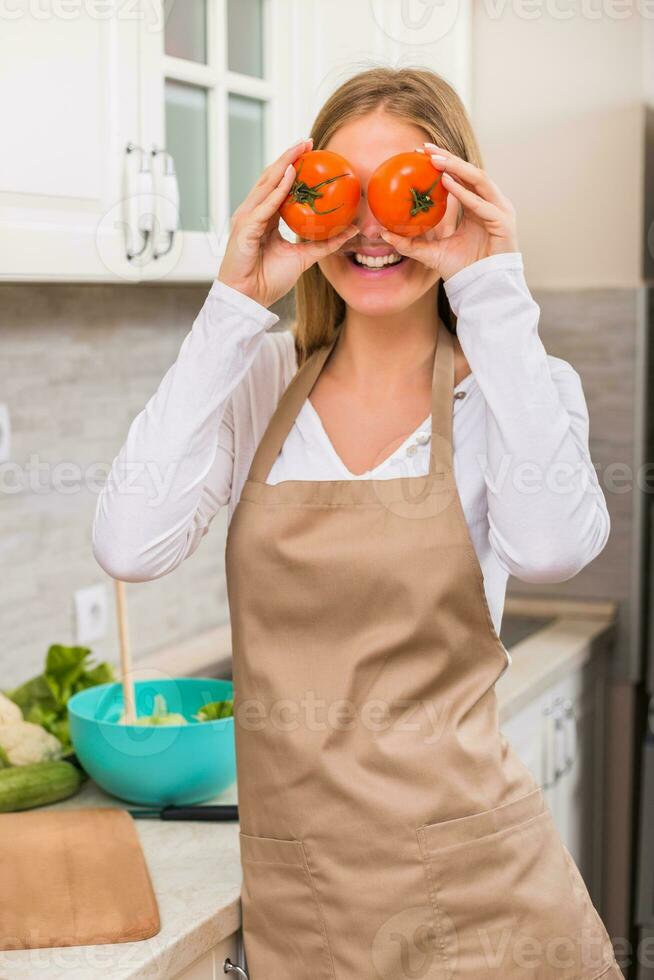 Beautiful woman covering eyes with tomato while making meal in her kitchen photo