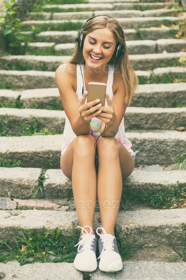 Beautiful young woman with headphones using mobile phone while sitting on the stairs in the city photo