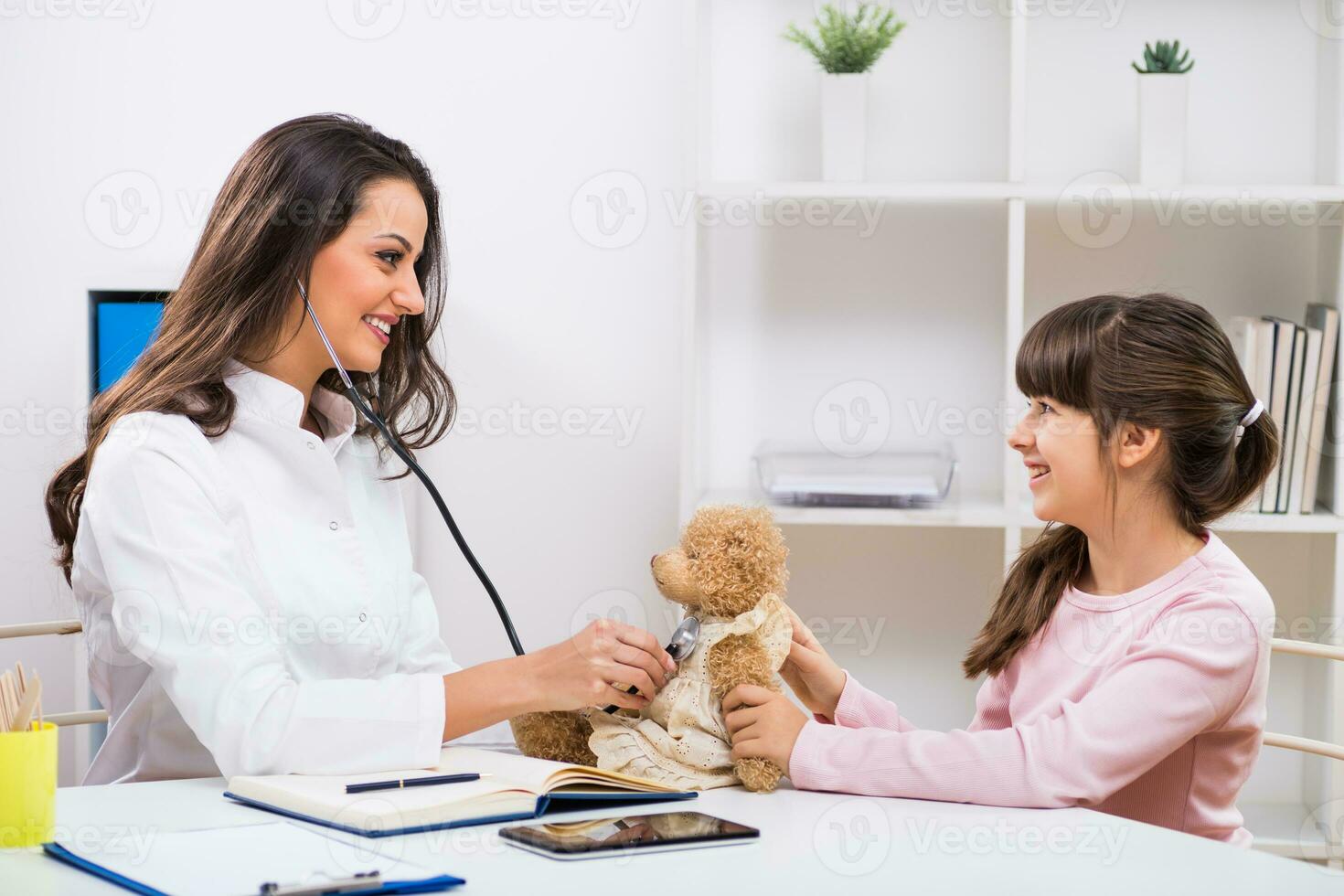 Female doctor and little girl examining teddy bear at medical office photo