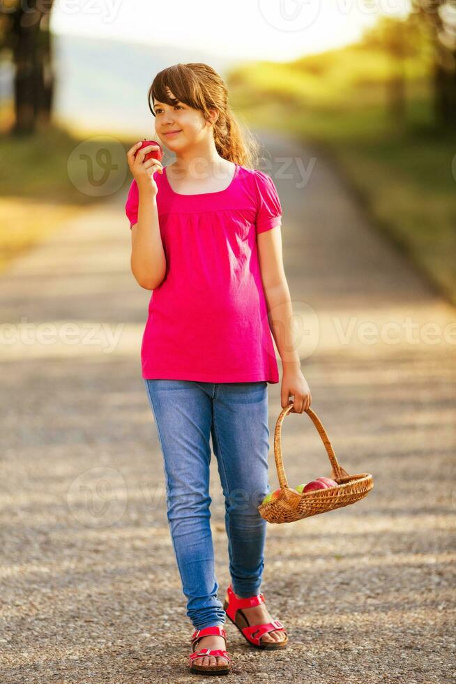 hermosa pequeño niña disfruta comiendo manzana mientras caminando en el naturaleza foto