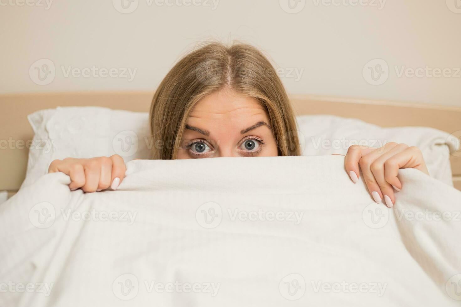 Woman with beautiful blue eyes laying in bed and peeking behind duvet photo