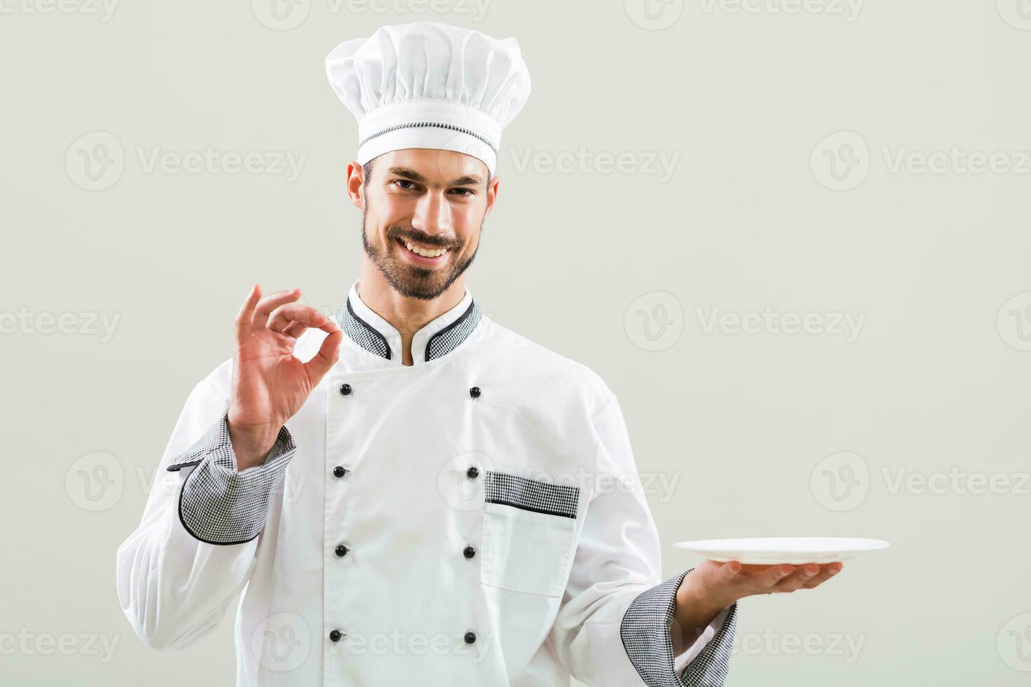 Smiling chef is holding plate and showing ok sign on gray background photo