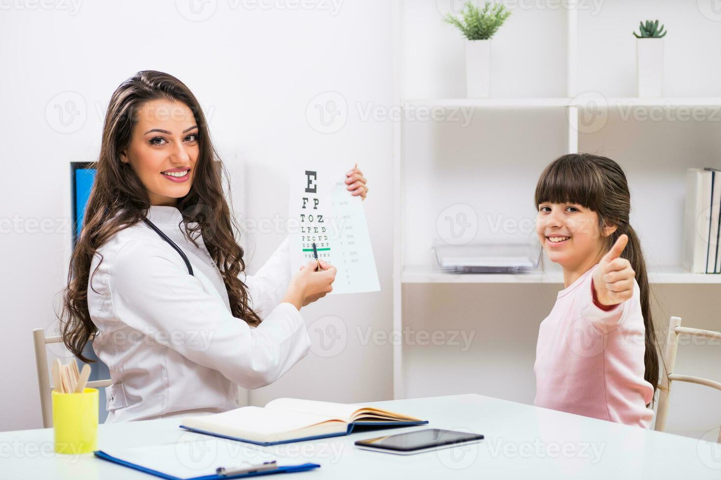 hembra médico haciendo ojo examen con niño a el médico oficina mientras pequeño niña demostración pulgar arriba foto