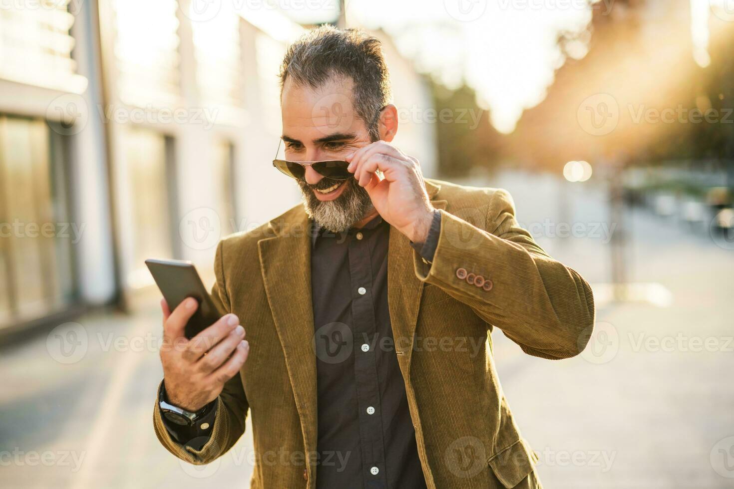 Modern businessman with beard using mobile phone while walking on the city street photo