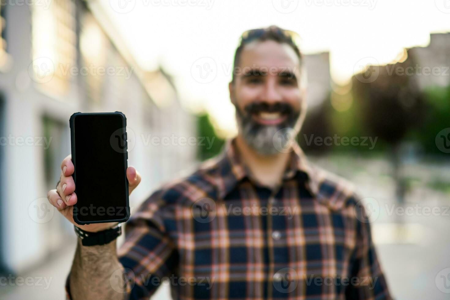 Modern businessman with beard showing blank screen of phone while standing on the city street. Focus on the phone. photo