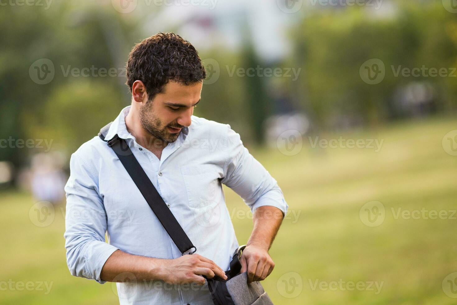 Young businessman standing at the park and searching something in his bag photo