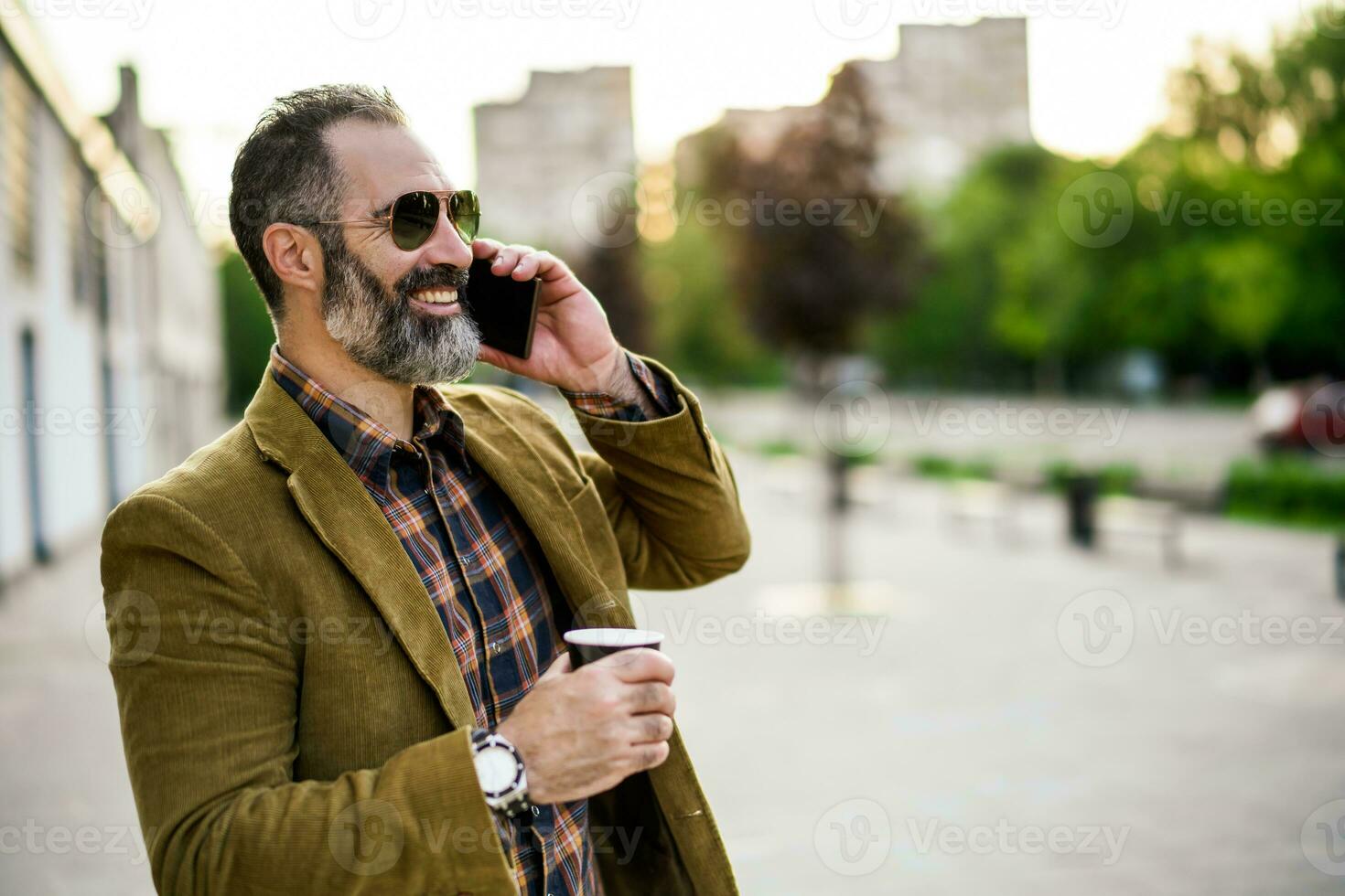 Portrait of modern businessman  with beard using mobile phone and drinking coffee while standing on the city street photo