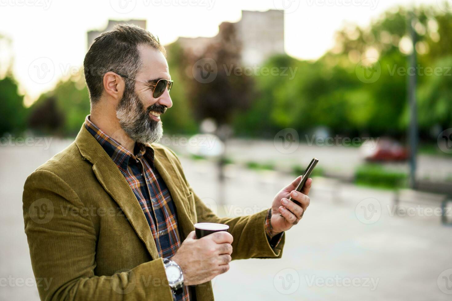 Portrait of modern businessman  with beard using mobile phone and drinking coffee while standing on the city street photo