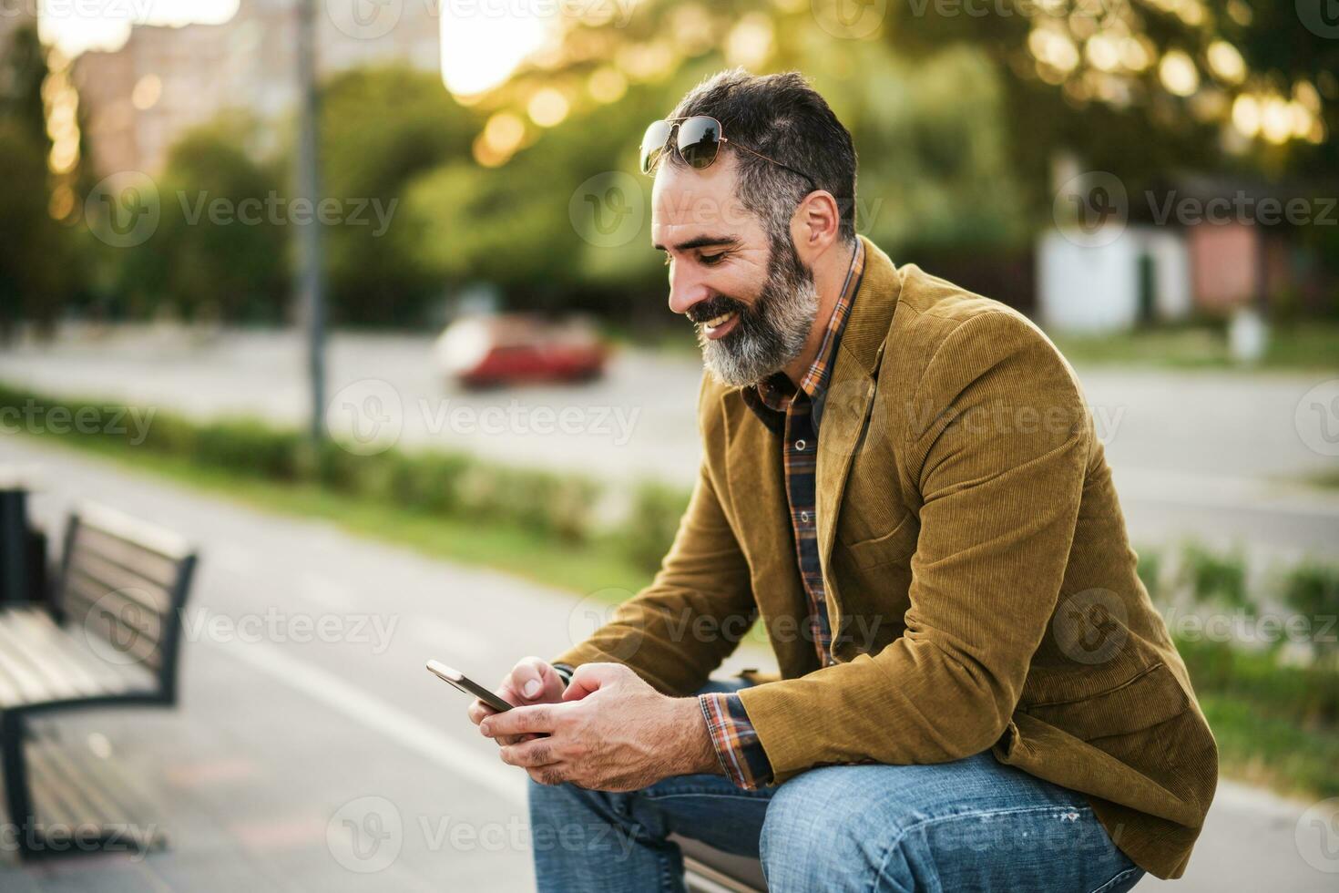 Modern businessman with beard using mobile phone while sitting on the bench in the city street photo