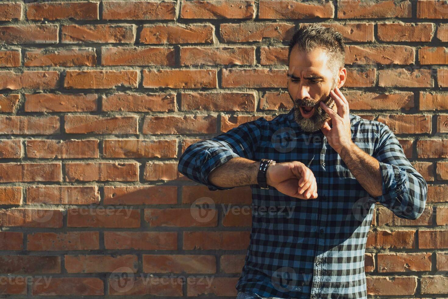 Businessman in panic with beard looking at watch while standing in front of brick wall outdoor photo