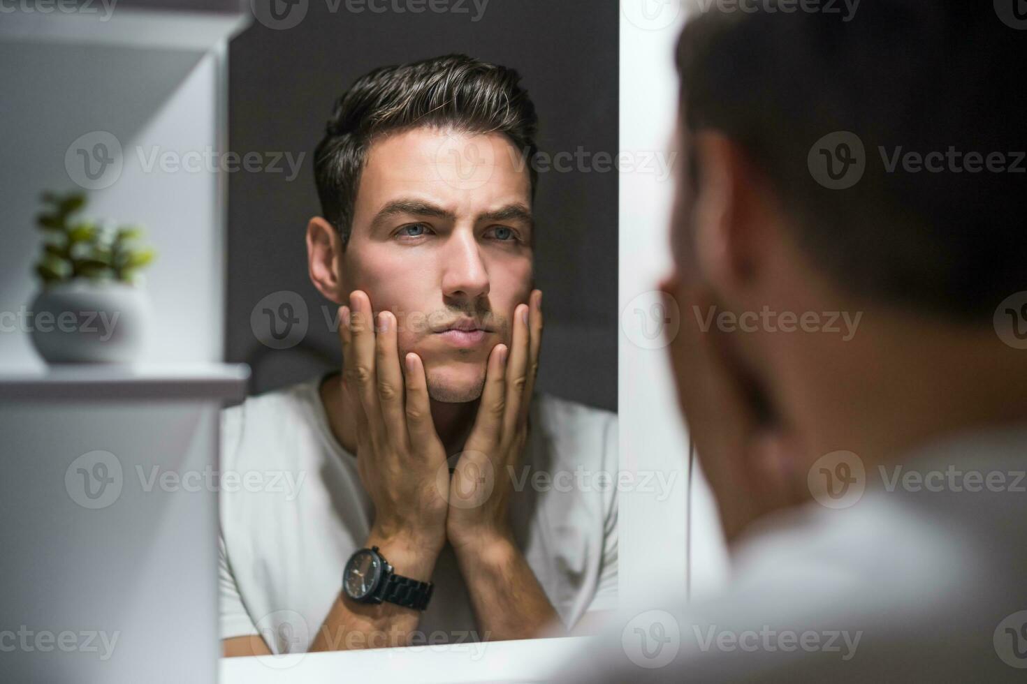 Tired man looking his eye bags and wrinkles while standing in front of mirror photo
