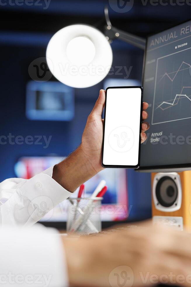 Detailed view of a male hand holding a mobile device with an isolated copyspace white screen in front of a desktop pc. Arab person displaying a blank chromakey mockup template on a smartphone. photo