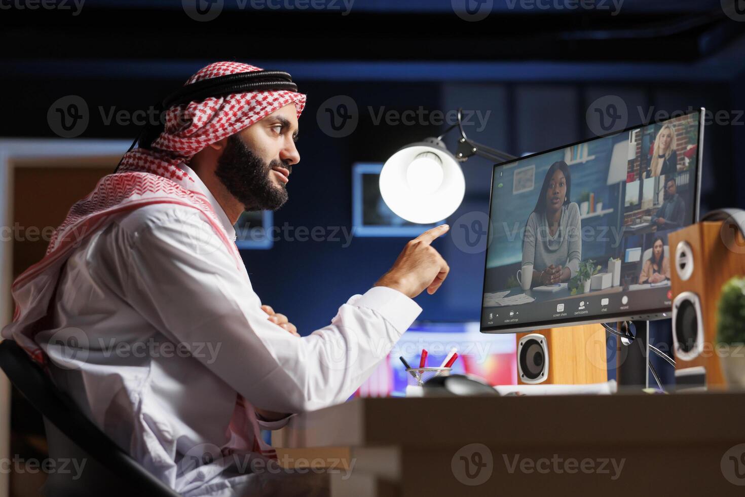 en computadora, musulmán chico es cuidadosamente escuchando a su compañeros de trabajo durante grupo vídeo conversación. vista lateral foto muestra un medio oriental hombre vestido tradicionalmente participación un en línea reunión.