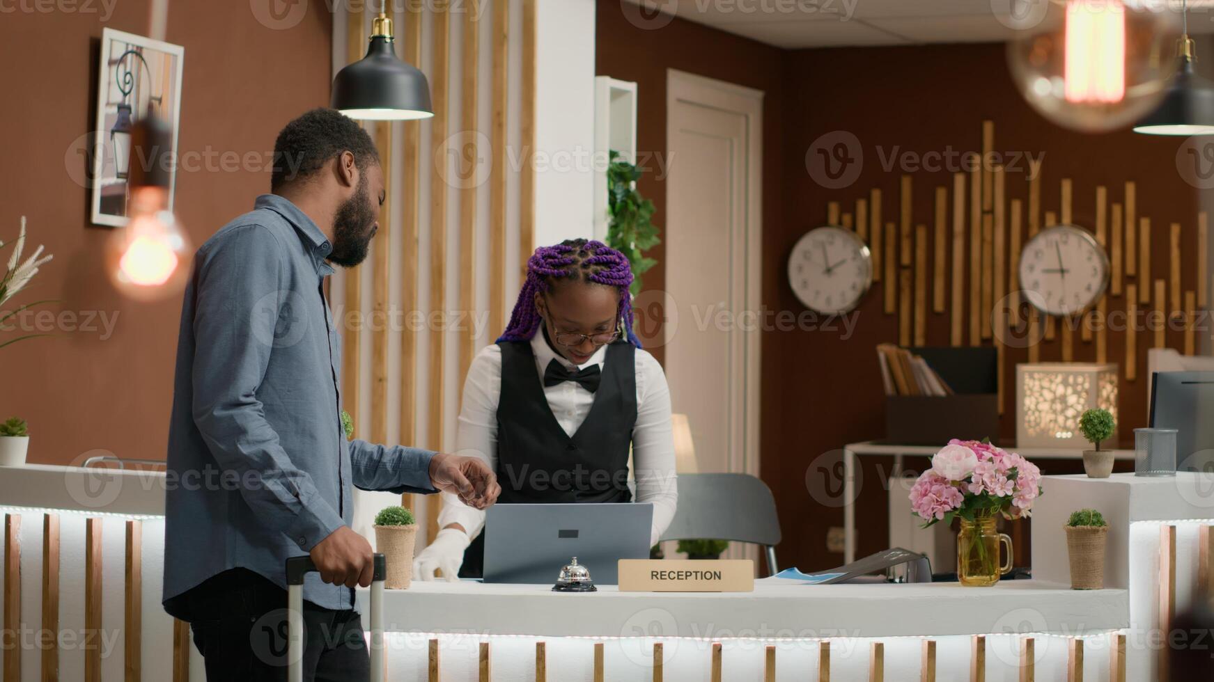 Person calls for service and room card at reception using front desk bell to talk to receptionist in lobby. African american hotel guest with trolley bags receiving access key for accommodation. photo