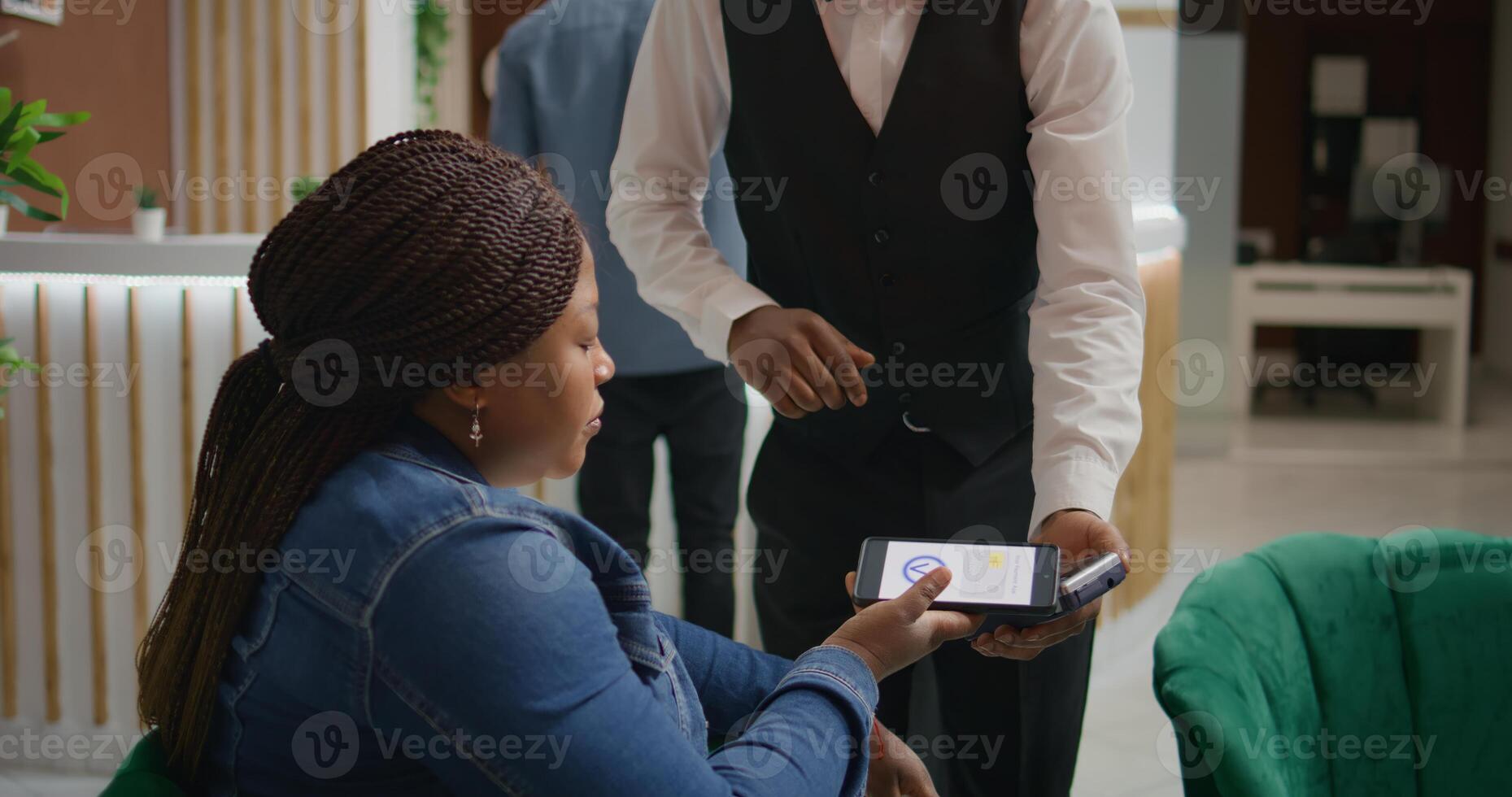 Woman does nfc mobile payment at pos to pay for cup of coffee while waiting on room reservation in hotel lounge area. African american tourist talking to porter about excellent concierge service. photo