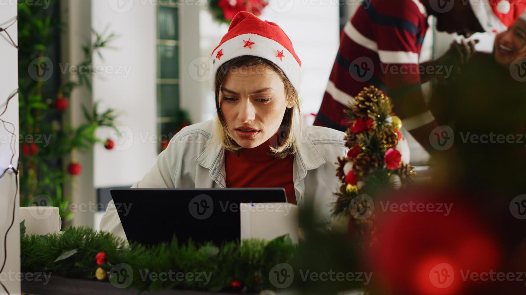 Woman filling paperwork in Christmas decorated office, perturbed by noisy coworkers discussing in background. Employee trying to concentrate on job while colleagues take shift break photo