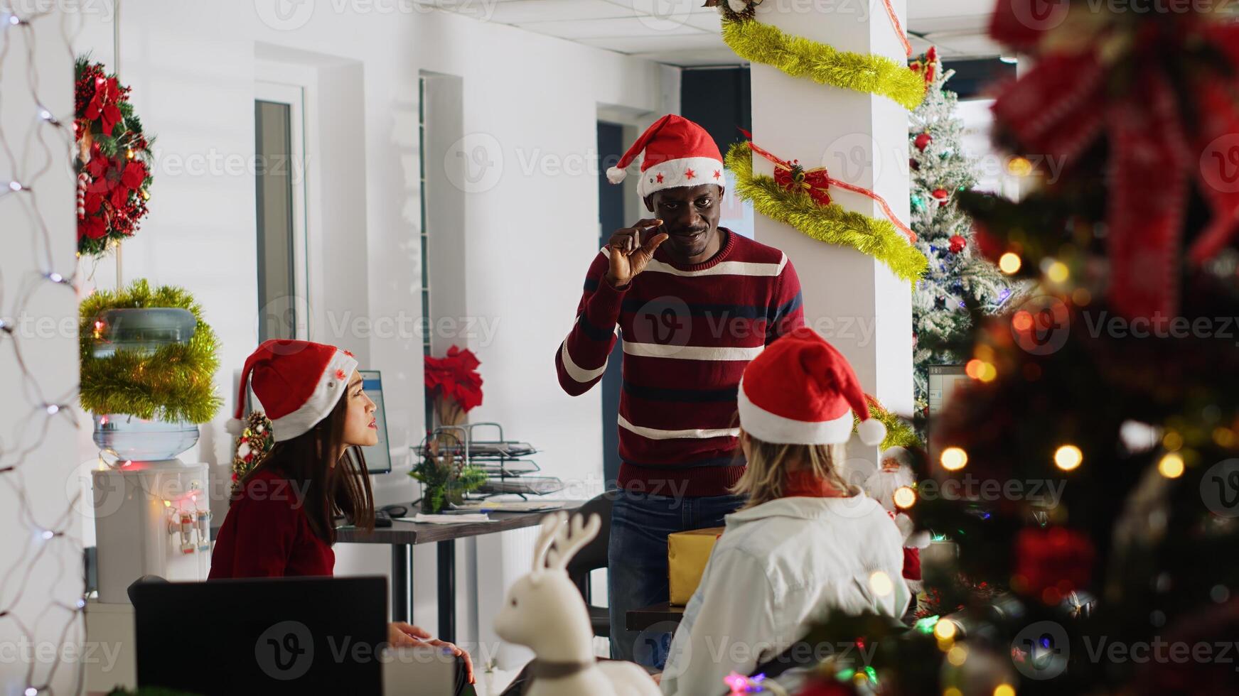 African american employee wearing Christmas hat entertaining coworkers by playing pretending game during break. Colleague having fun doing pantomime during Secret Santa party photo
